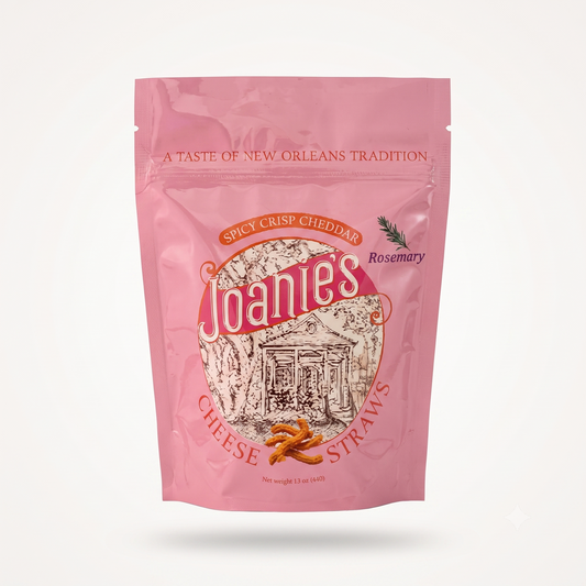 A pink stand-up pouch of Joanie's Spicy Crisp Cheddar Cheese Straws on a clean white background. The packaging features a prominent circular logo with the brand name 'Joanie's' in white script, an illustration of a New Orleans street scene, and small images of cheese sticks and rosemary.